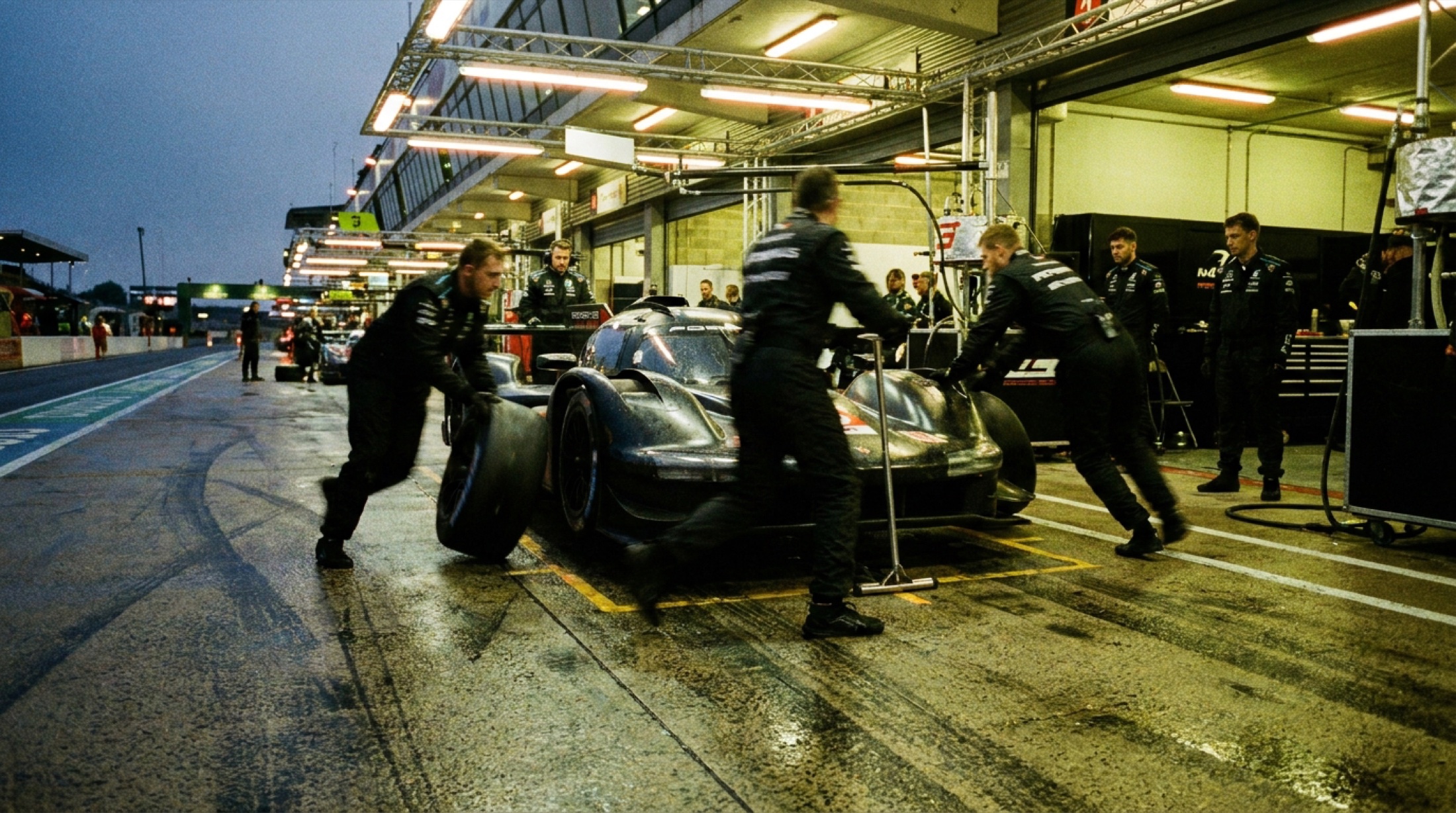 Le Mans pit şeridinde gece çalışması, mekanik ekip hareket halinde, ıslak asfalt ışıkları yansıtıyor
