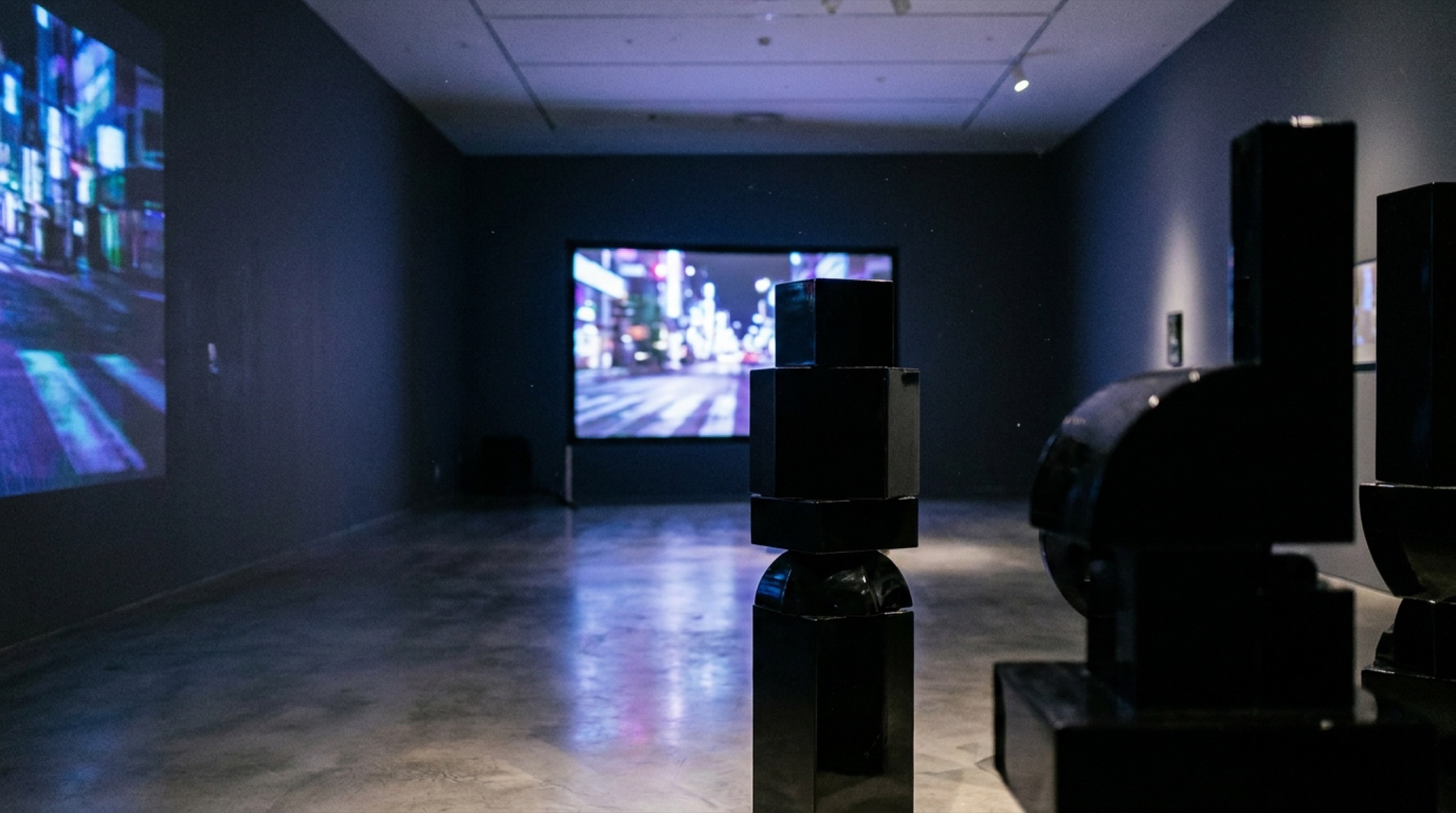 Kunstmuseum Basel'de video projeksiyonun mavi-mor ışığıyla aydınlanan karanlık çağdaş galeri; arka planda dijital kentsel manzara görüntüsü.