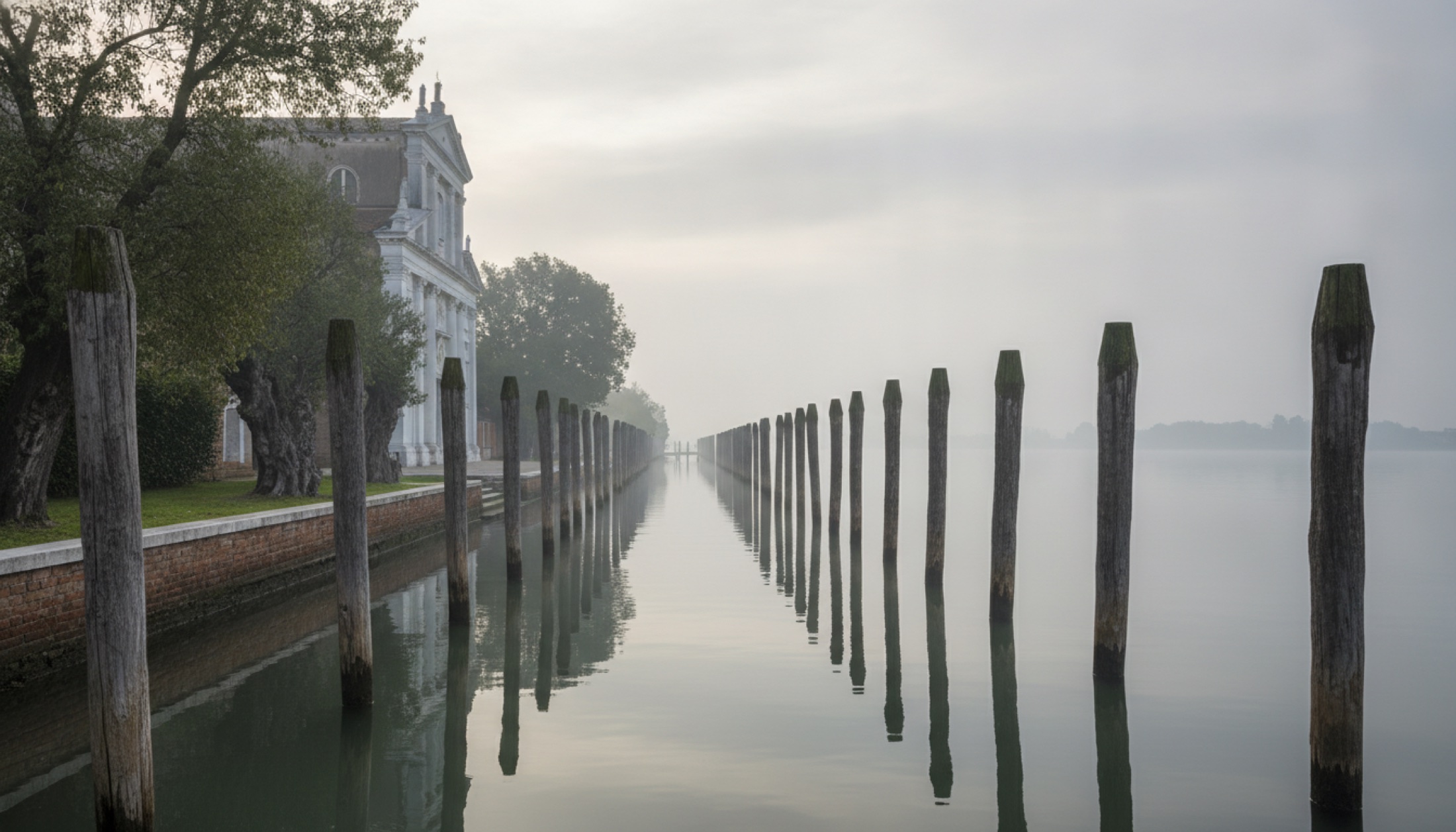 Venedik lagununda Torcello adasında Santa Maria Assunta Katedrali cephesi ve önünde ahşap kazıklarıyla sakin bir kanal, sabah sisinin içinde.