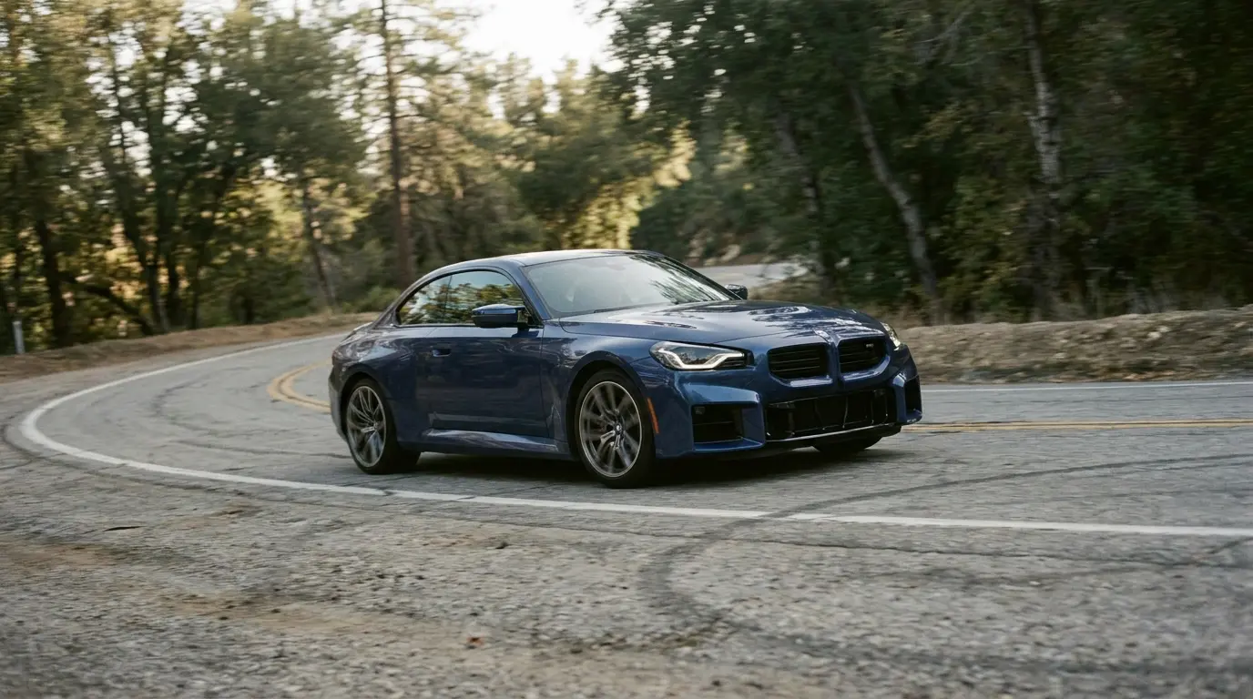 2026 BMW M2 xDrive G87, kavisli bir dağ yolunda, arka planda çam ağaçları ve bulutlu gökyüzü
