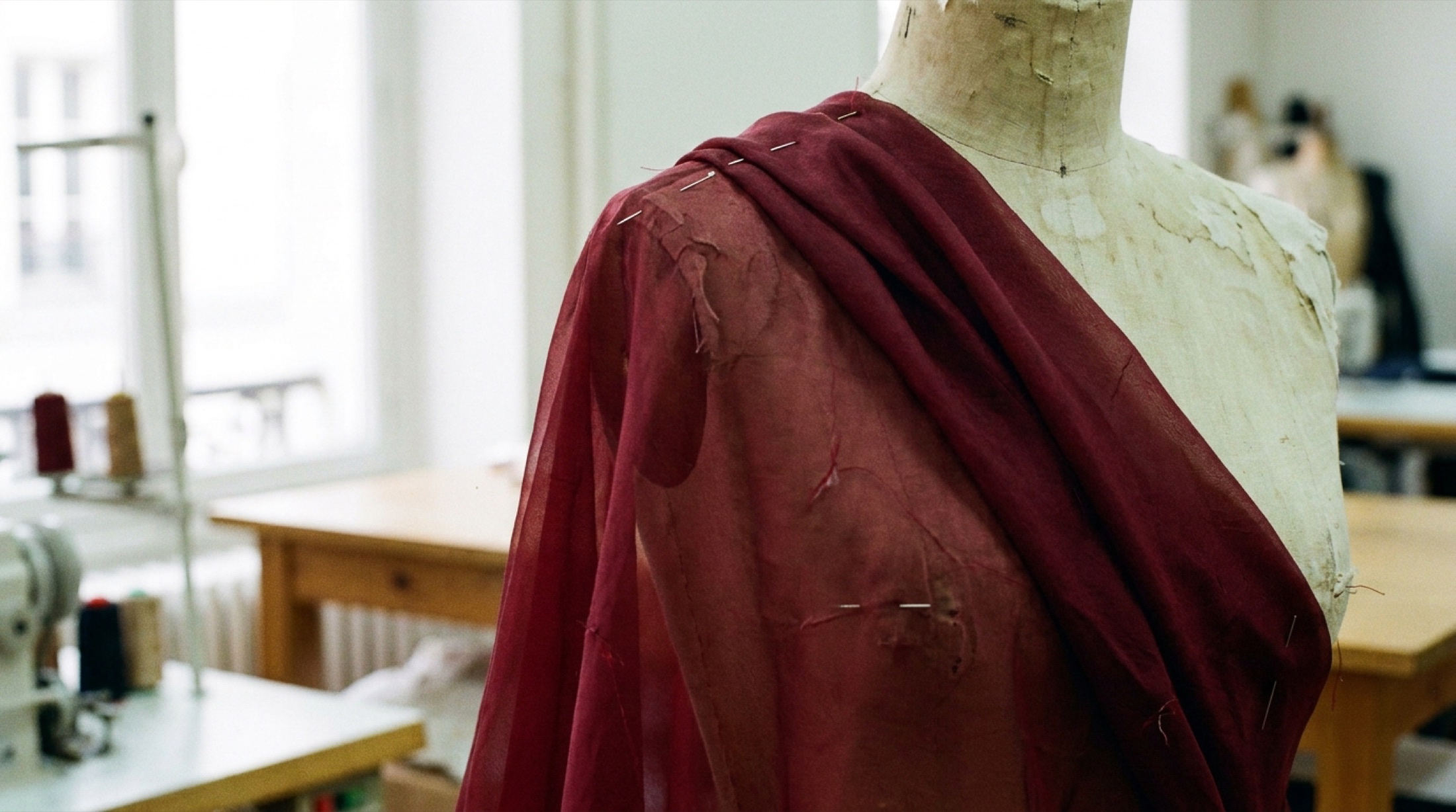 Atölyede manken üzerine pimlerle tutturulmuş bordo ipek organza drapesi, sol pencere ışığı