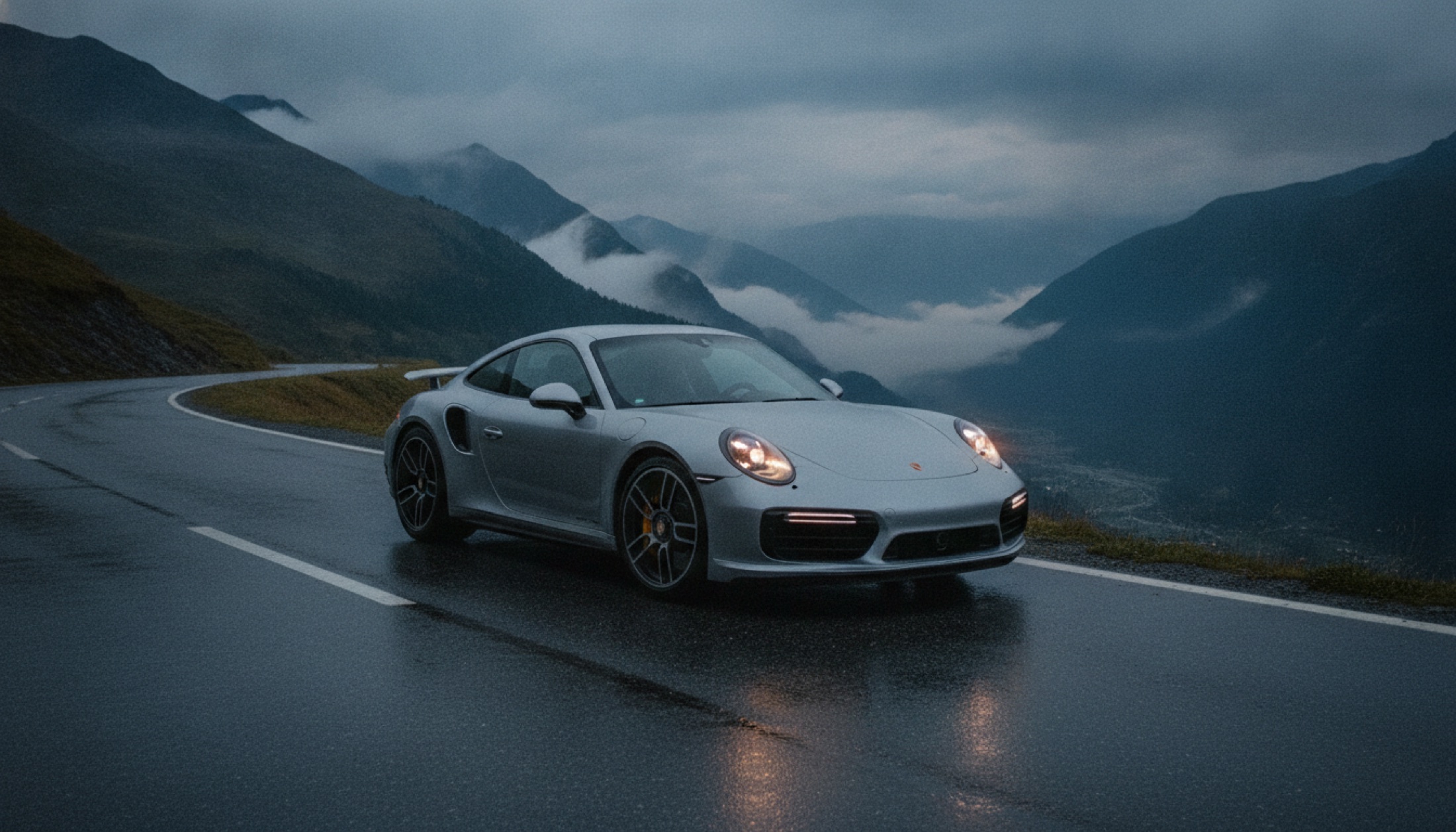 Alp dağlarında ıslak yolda duran Porsche 911 Turbo S, aktif arka spoyleri açık, gün batımı ışığı