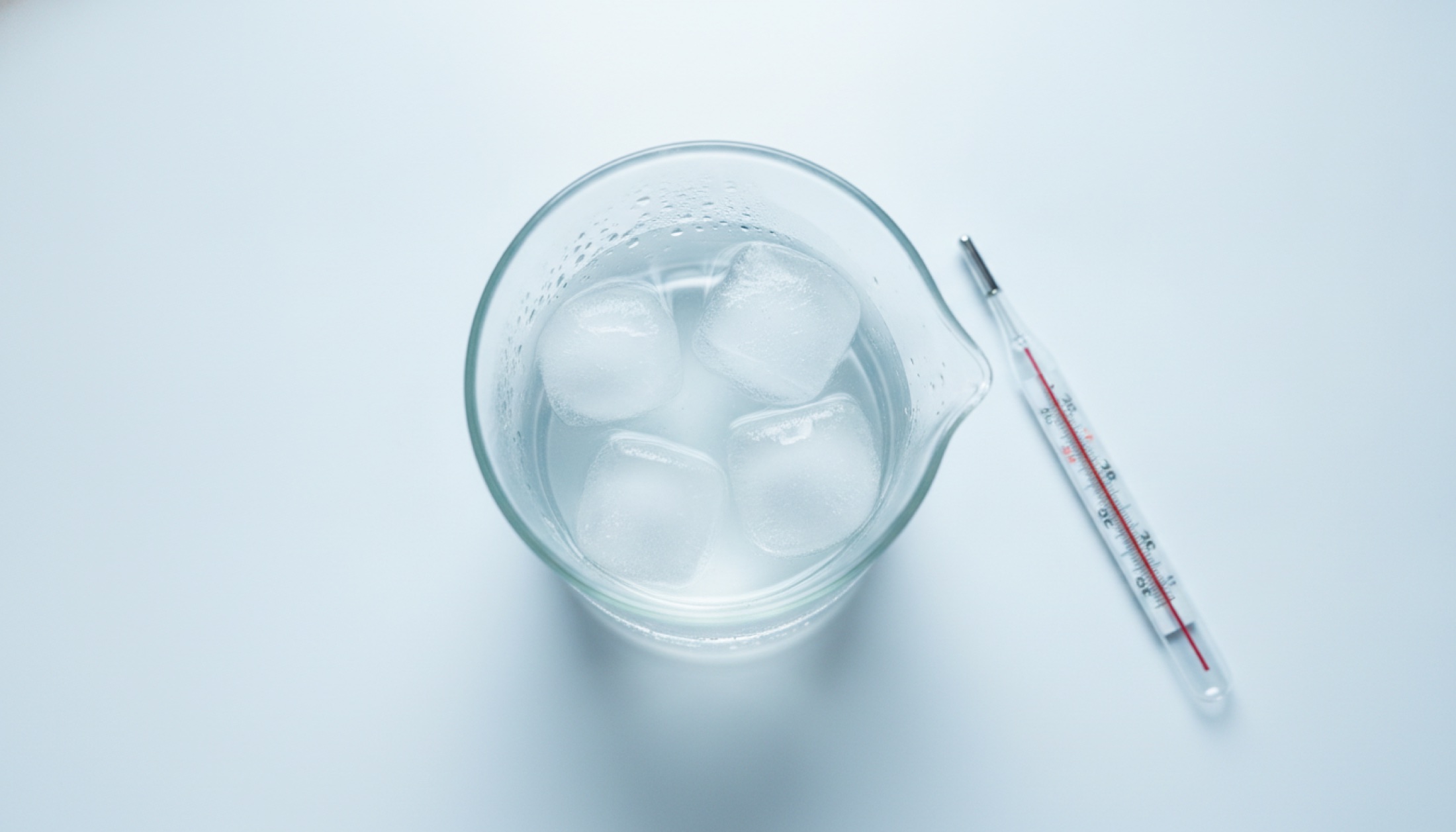 Buz dolu bir cam beher ve yanında termometre — soğuk su terapisinin bilimsel incelemesini simgeleyen klinik bir still-life.