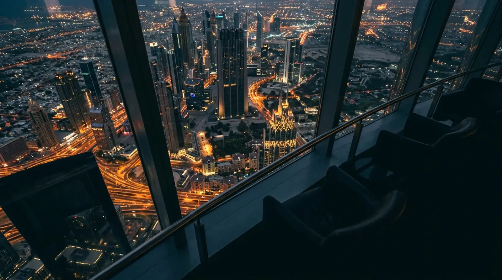 Gece vakti cam cepheli yüksek binanın içinden dışarıya bakan geniş açılı fotoğraf; altta şehir ışıkları, üstte bulutlu gökyüzü, ön planda derin gölgeler